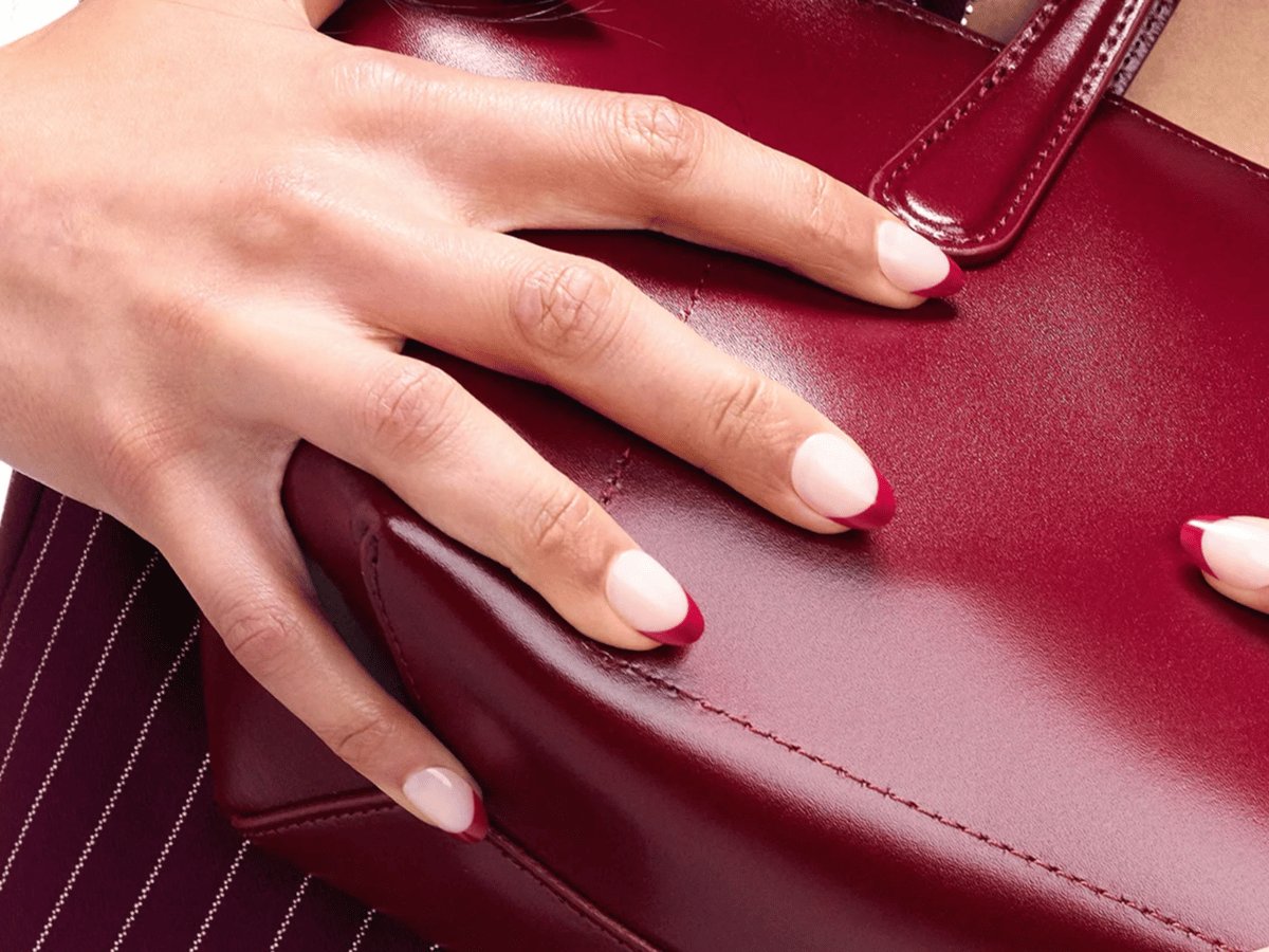 Close-up of a hand with blush pink press-on nails featuring deep red French tips, resting on a burgundy leather handbag